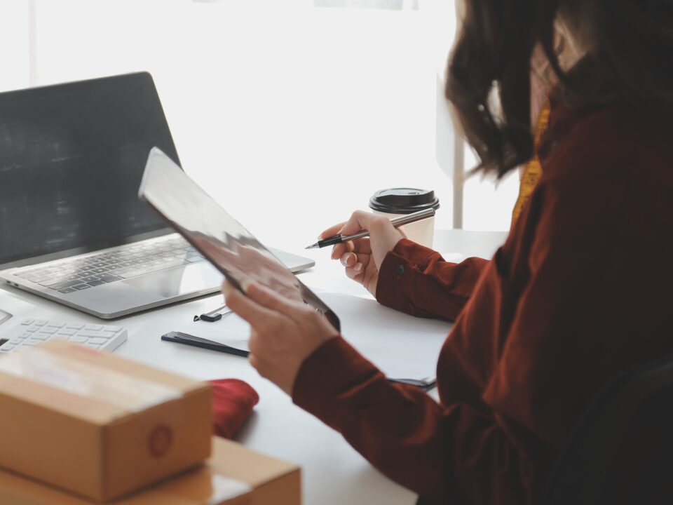 Eine Person sitzt an einem Tisch vor einem Laptop und hält in der rechten Hand eine Kugelschreiben, um etwas zu notieren und in der linken Hand hält sie ein Tablet