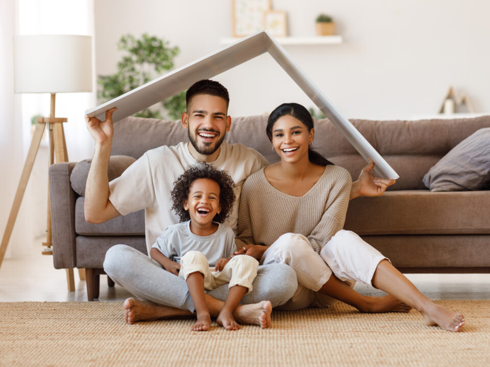 Ein Elternpaar sitzt mit ihrem kleinen Sohn vor einem Sofa und der Vater animiert mit zwei Bretten ein Dach über deren Kopf.