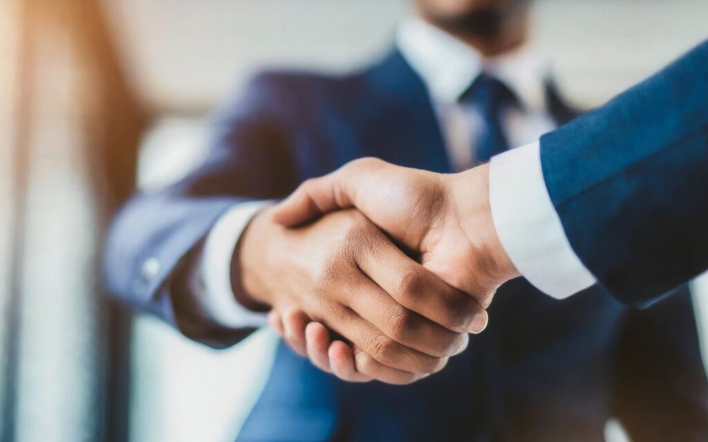 Nahaufnahme von zwei Personen in Anzügen, die sich die Hände schütteln und damit eine geschäftliche Vereinbarung oder Partnerschaft symbolisieren. Der Fokus liegt auf ihren Händen, mit unscharfem Hintergrund, wodurch eine professionelle und einladende Atmosphäre entsteht, die die Fünftelregelung bei Abfindungen widerspiegelt.