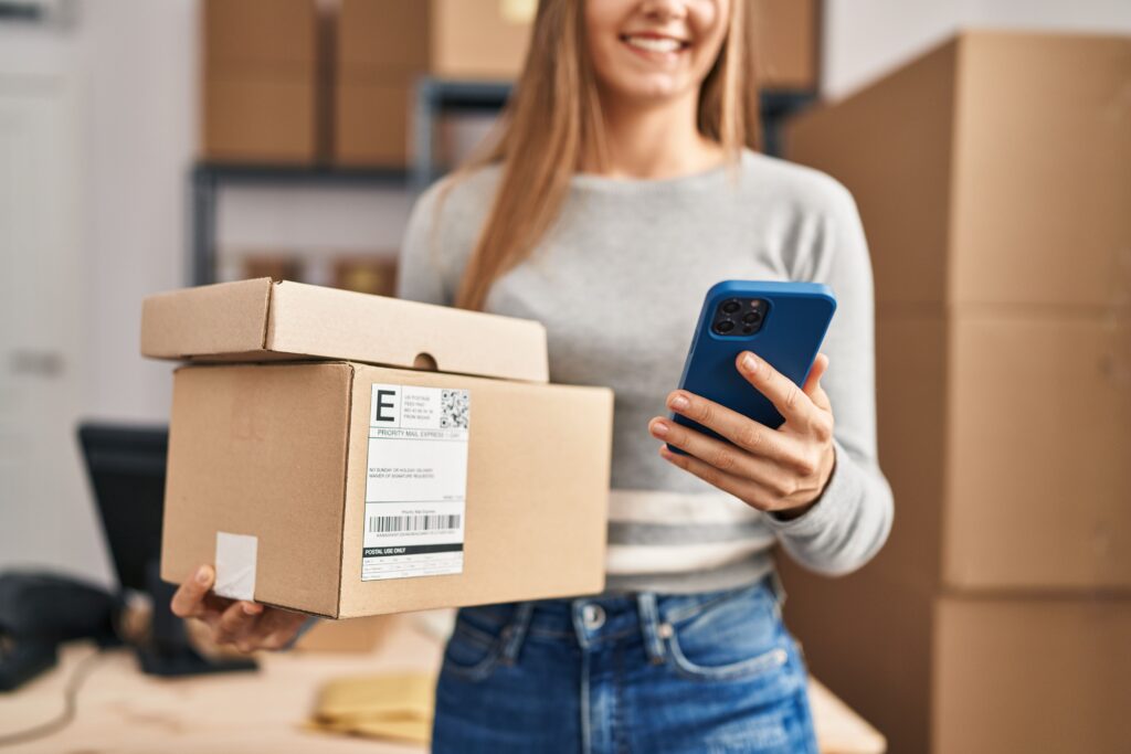 Frau mit Karton und Smartphone lächelt in einem Raum voller Regale mit Boxen, wirkt wie ein Versandbereich.