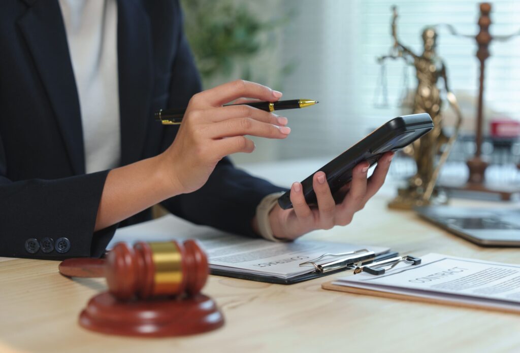 Person im Anzug am Schreibtisch mit Smartphone und Stift, umgeben von Richterhammer, Waage, Vertragsdokumenten und Laptop. Sie rechnet die Erbschaftssteuer für Geschwister aus
