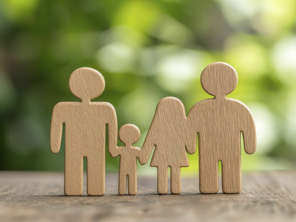 Hölzerne Familienfiguren mit vier Personen, die sich an den Händen halten, auf einer Oberfläche mit einem verschwommenen grünen Hintergrund, der an einen Garten erinnert.