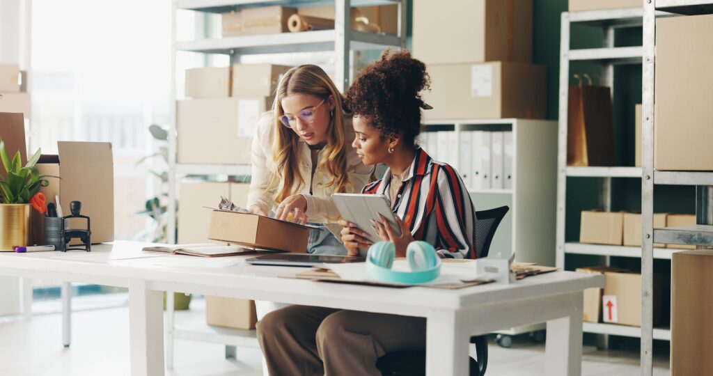 Zwei Frauen arbeiten zusammen in einem Büro mit Kartons und benutzen ein Tablet und ein Klemmbrett für den Versand oder die Bestandsverwaltung.