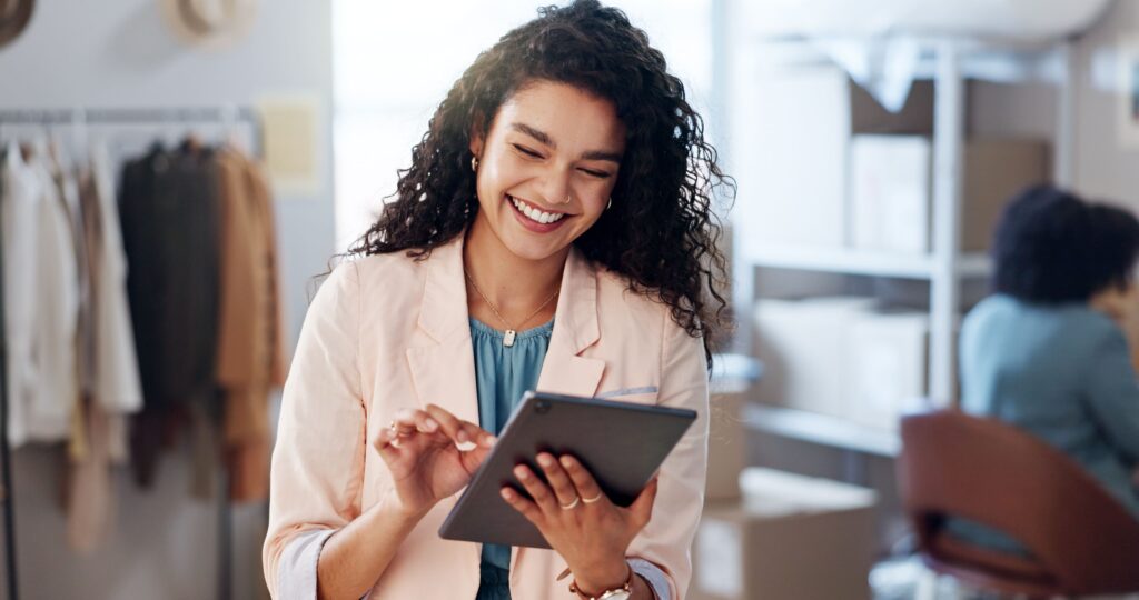 Lächelnde Frau mit lockigem Haar in einem rosa Blazer benutzt ein Tablet in einem hellen Büro mit Regalen im Hintergrund.
