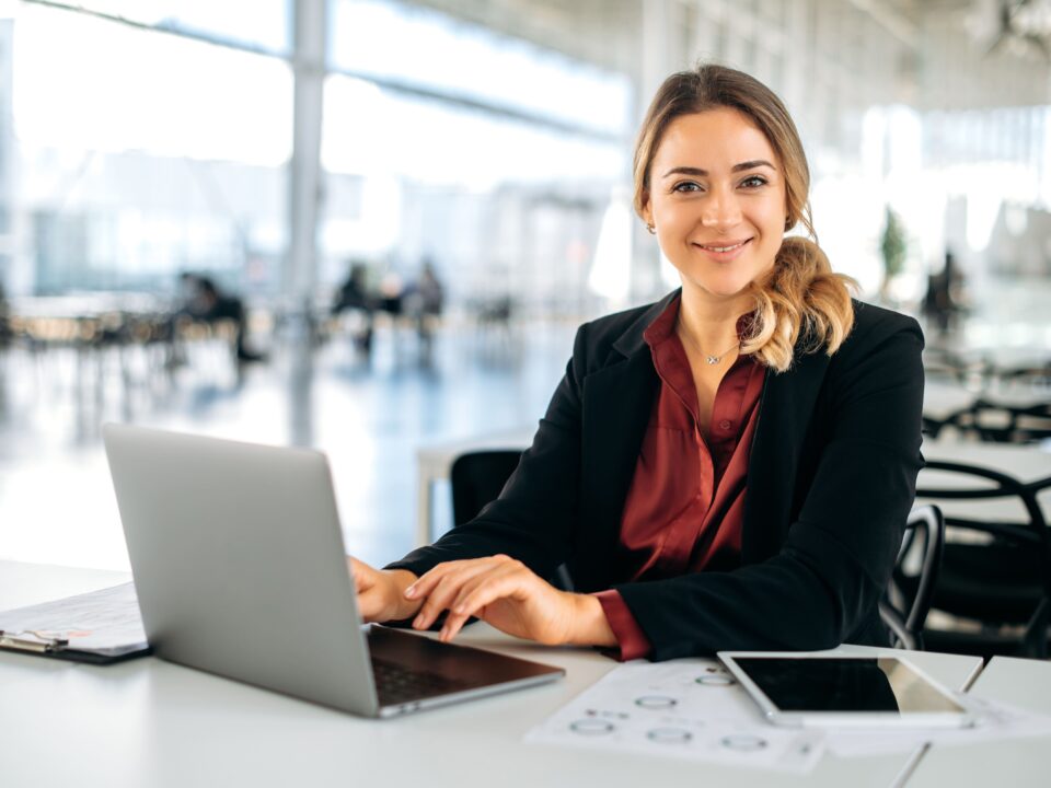 Geschäftsfrau lächelt am Schreibtisch mit Laptop, Tablet und E-Commerce-Dokumenten in einem hellen Büro mit großen Fenstern.