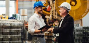Zwei Männer mit Schutzhelmen schütteln sich in einer Industrieumgebung die Hand, einer hält ein Tablet.
