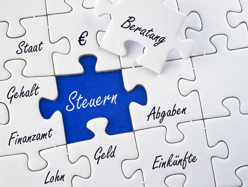 Bild von einem Puzzle, auf dem im Mittelpunkt blau hinterlegt “Steuern” steht und daneben Begriffe wie Einkünfte, Geld und Finanzamt sowie auf einem herausgelösten Puzzleteil “Beratung”.